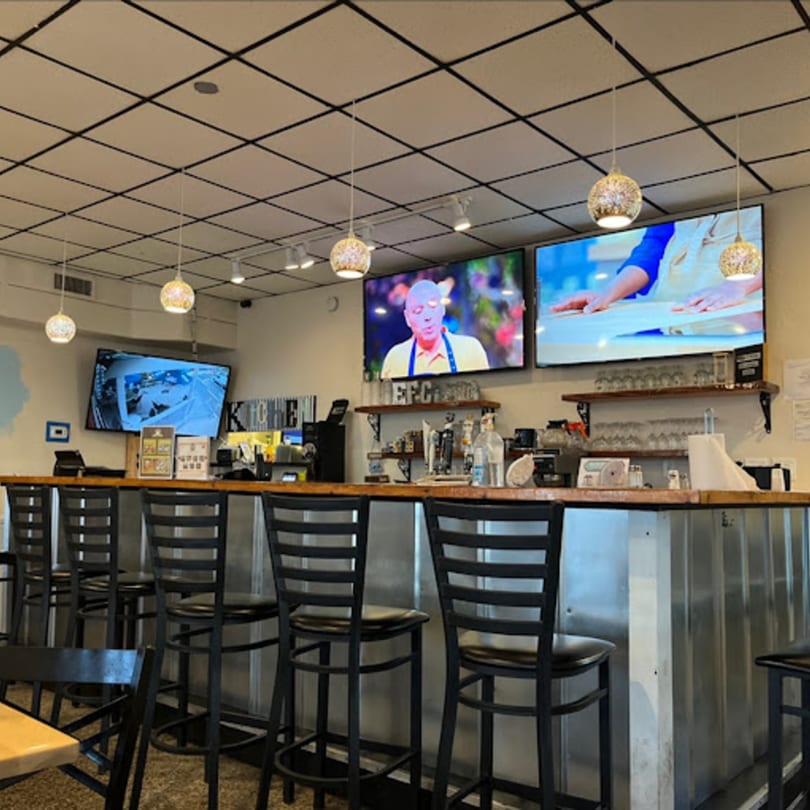 Interior of Gili's Kitchen showing a bar with stools, shelves, and multiple TVs displaying content.