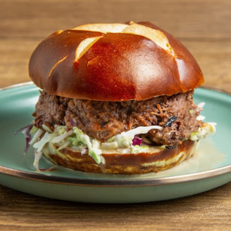 A Gili's Kitchen kosher pulled beef sandwich with coleslaw on a pretzel bun, served on a teal plate.