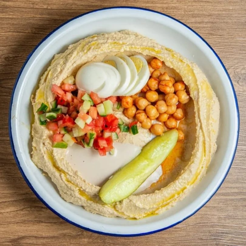 A bowl of Gili's Kitchen hummus topped with chickpeas, diced vegetables, sliced egg, and a pickle.