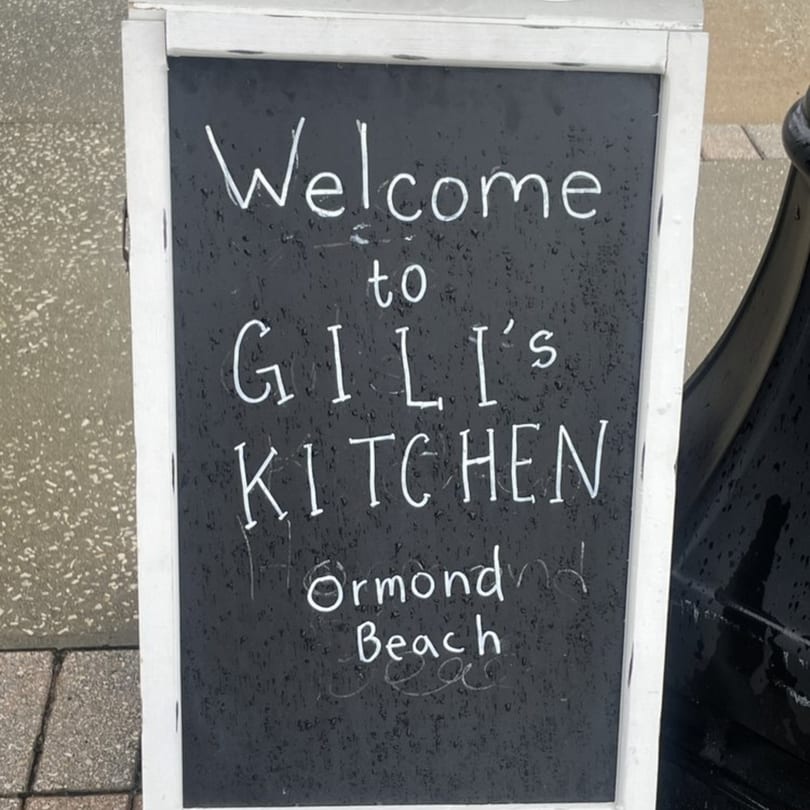 A chalkboard sign with "Welcome to Gili's Kitchen Ormond Beach" written in white chalk.