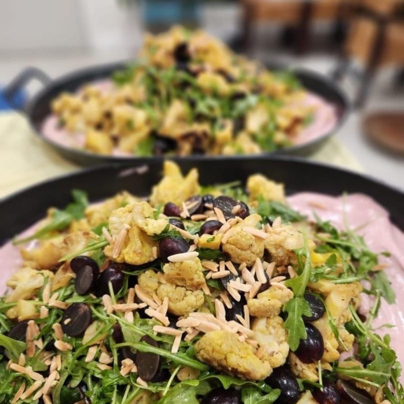 Two pans of roasted cauliflower and black olives on a bed of arugula with pink sauce.