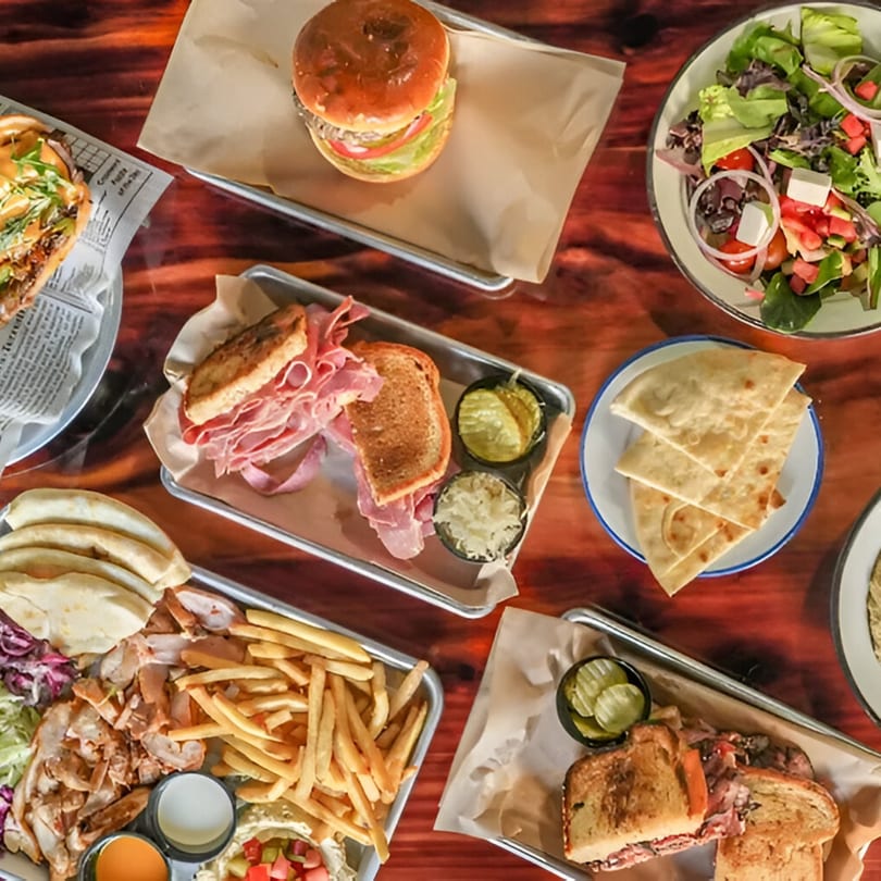 Overhead view of various kosher dishes including sandwiches, fries, salad, and pita bread on a wooden table.