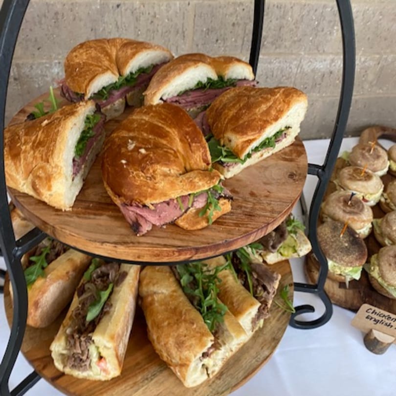 Tiered wooden trays display various sandwiches, including beef and arugula on croissants and baguettes.